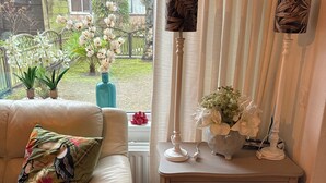 Een woonkamer met een beige bank, een witte bijzettafel met een lamp en een vaas met bloemen.