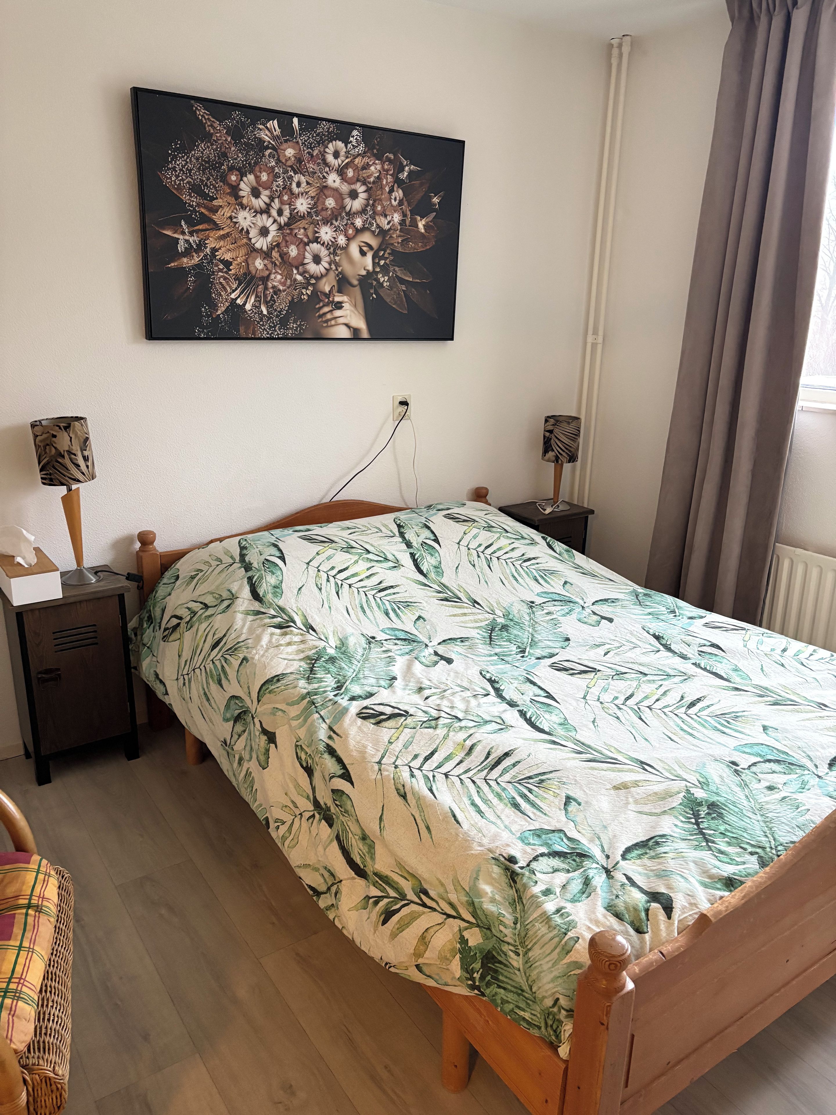 Un dormitorio con cama, mesita de noche, lámpara, un cuadro colgado en la pared y ventana con cortinas.