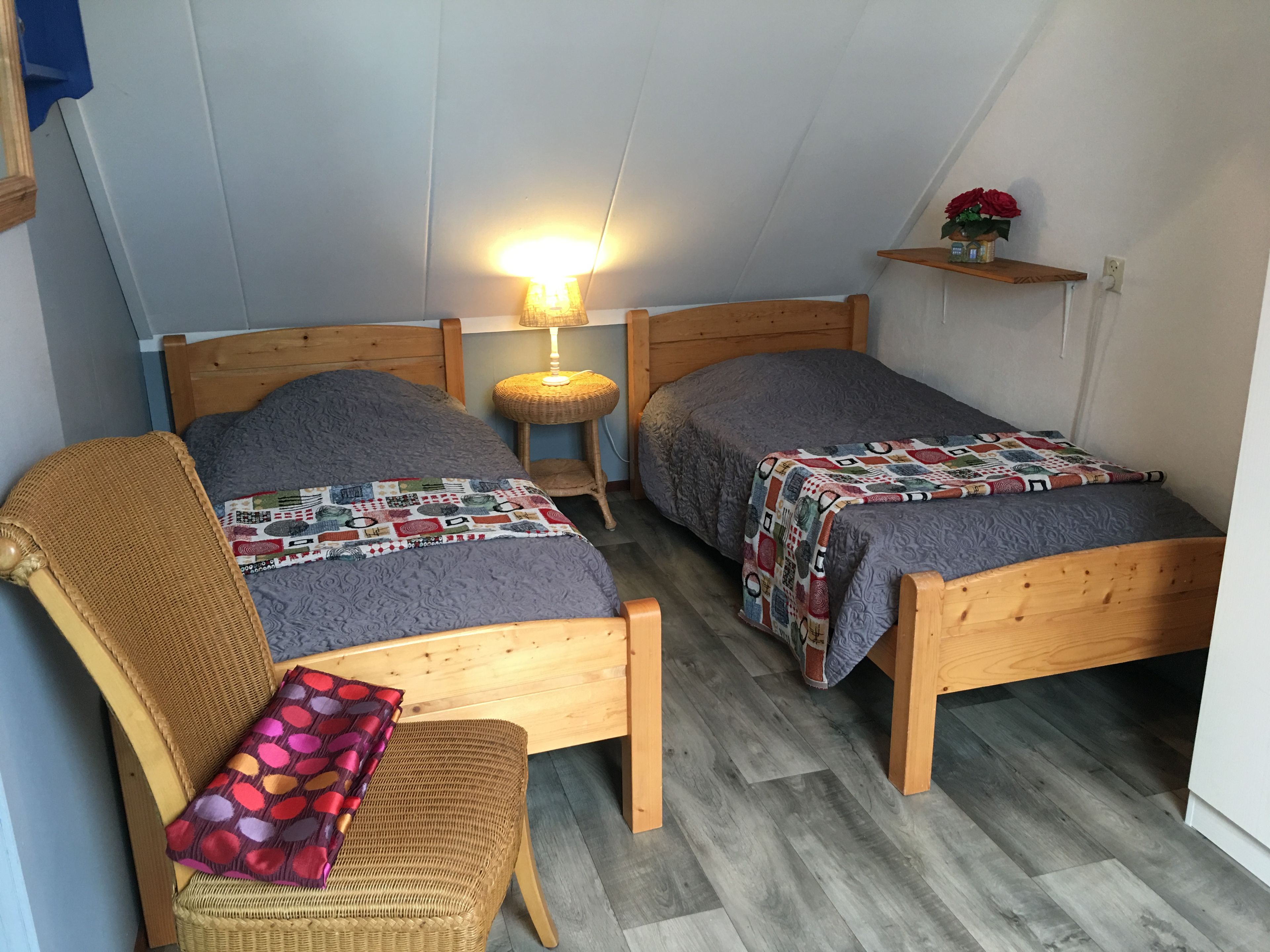 A small, cozy room with two wooden beds, a wicker chair, and a nightstand with a lamp.