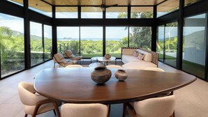 A spacious room with a large round table, comfortable seating, and large windows offering a view of lush greenery and hills. - Casa Armonía - New Luxury Ocean View Villa - Walking distance to the beach! (Playa Hermosa)