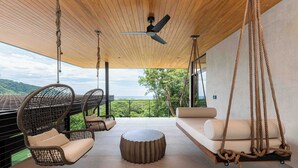 A modern outdoor seating area with wicker chairs, a sofa, and a swing chair, overlooking a lush landscape. - Casa Armonía - New Luxury Ocean View Villa - Walking distance to the beach! (Playa Hermosa)