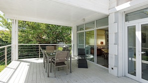 Una terraza cubierta al aire libre con una mesa y sillas, una puerta de cristal que da al interior y una vista a la vegetación de fondo.