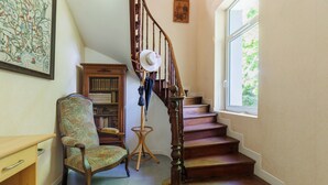 Una escalera con escalones de madera y barandilla, una silla con cojín estampado, una pequeña mesa de madera y una ventana con vistas a follaje.