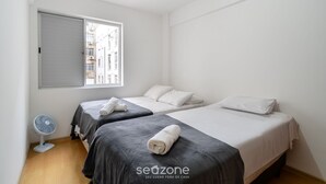 Ein aufgeräumter, minimalistischer Raum mit zwei Betten, einem Standventilator und einem Fenster mit Blick auf Gebäude.