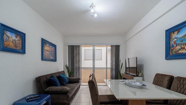 Ein modernes Wohnzimmer mit einem braunen Sofa, einem weißen Esstisch und einem Fernseher. An den Wänden hängen zwei gerahmte Bilder und ein Fenster mit Vorhängen.