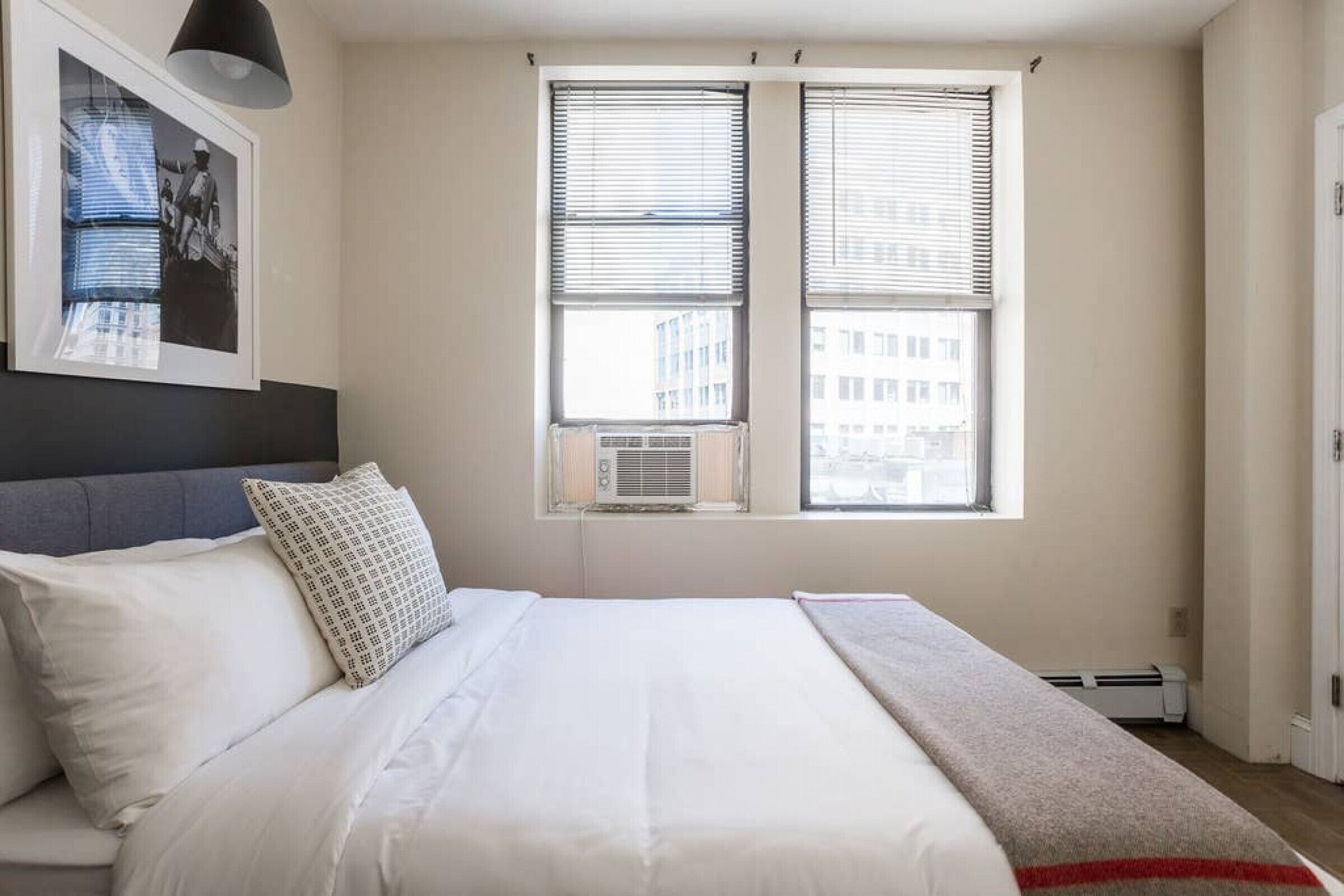 A bedroom with a bed, two windows with blinds, an air conditioning unit, and a framed picture on the wall.