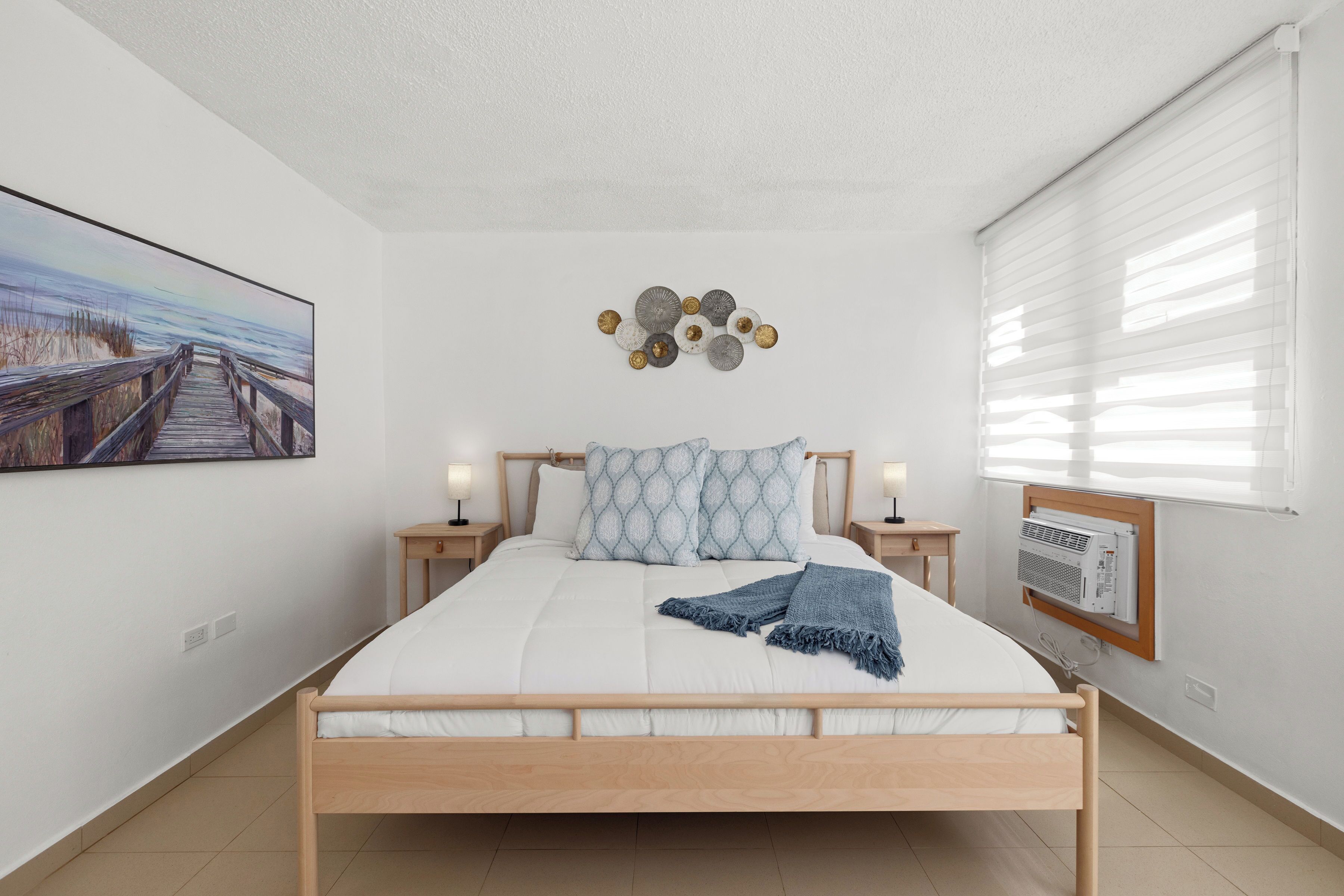 A bedroom with a wooden bed, white bedding, a blue throw blanket, a wall-mounted air conditioner, and a framed picture of a beach scene.