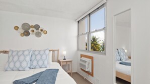 Une chambre à coucher comprenant un lit, une table de chevet, une lampe, des œuvres d’art murales et une fenêtre donnant sur des palmiers.