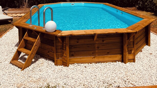 Une piscine hors sol en bois, dotée d’un escalier, entourée de galets blancs et de verdure.