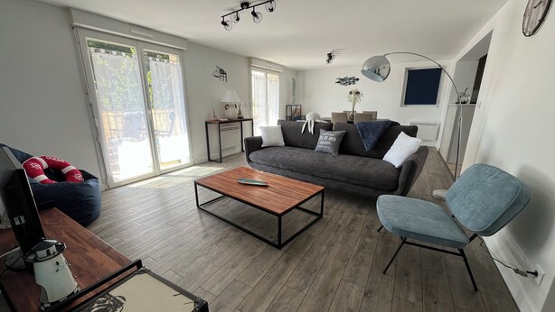 A modern living room with a grey sofa, a wooden coffee table, and a blue armchair. There is a lamp, a clock on the wall, and a sliding glass door leading outside. - Le Rameau, gite in the heart of the city, terrace, quiet street close to the Old Port. (Pornic)