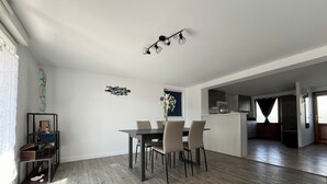 A modern dining area with a wooden floor, a central table, and four chairs. There is a kitchen area in the background with a window and a door. - Le Rameau, gite in the heart of the city, terrace, quiet street close to the Old Port. (Pornic)