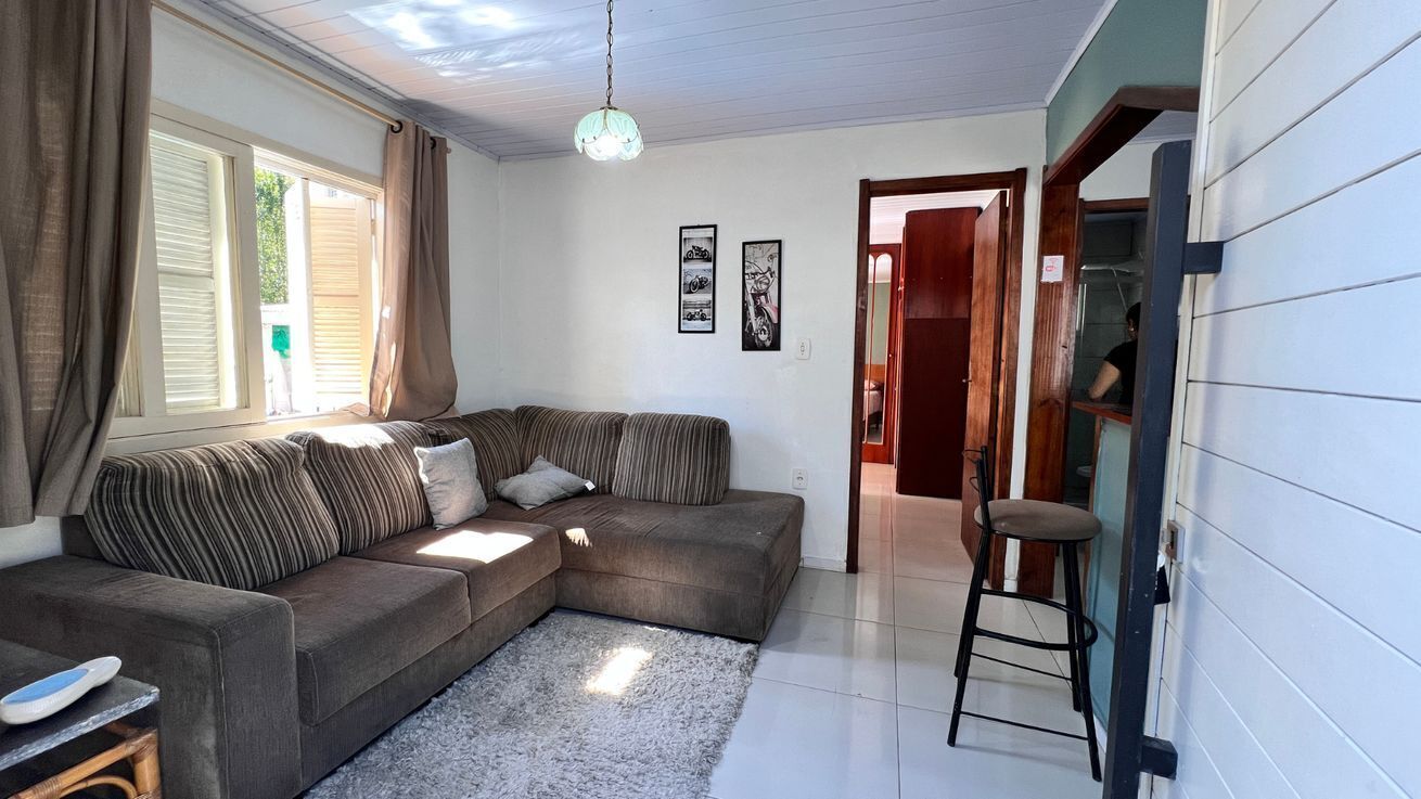 A living room with a sectional sofa, a small table, and a chair. There are two framed pictures on the wall, a window with curtains, and a door leading to another room.