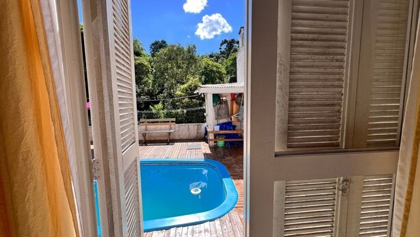 A view of a swimming pool with a wooden deck, a covered area with a table and chairs, and a clear blue sky with clouds. - Casa com Piscina, Churrasqueira e Mesa de Bilhar (Caxias do Sul)