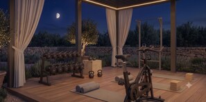 An outdoor gym area with exercise equipment, including dumbbells, kettlebells, and a stationary bike, set against a backdrop of trees and a stone wall. - VedoMare Hotel & Suites (Zakynthos)