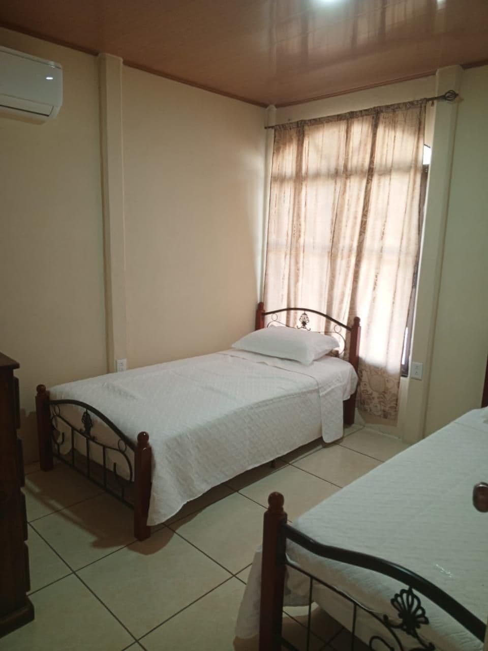 Una habitación pequeña con cama individual, estructura de madera, mesita de noche y ventana con cortinas.
