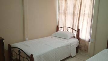 Una habitación pequeña con cama individual, estructura de madera, mesita de noche y ventana con cortinas.