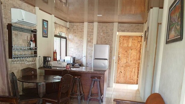 A spacious living area with a wooden sofa, a dining table with chairs, and a kitchen area with a refrigerator and bar shelves. - Casa Vacacional (Ciudad Cortes)