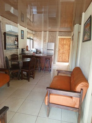 A spacious living area with a wooden sofa, a dining table with chairs, and a kitchen area with a refrigerator and bar shelves. - Casa Vacacional (Ciudad Cortes)