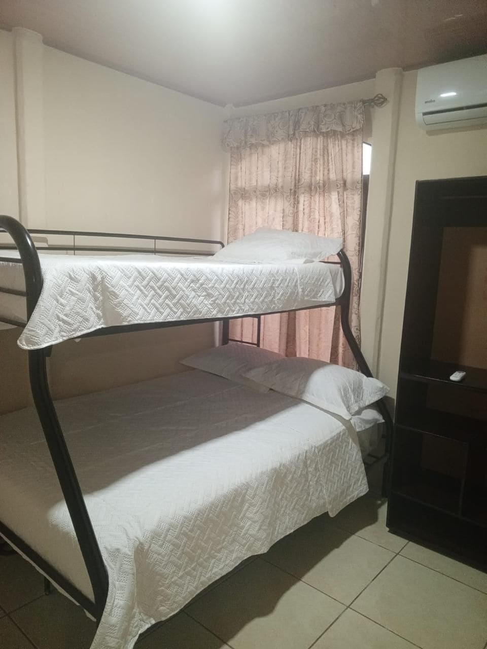 A bunk bed with a white bedspread, a window with curtains, and an air conditioning unit on the wall.