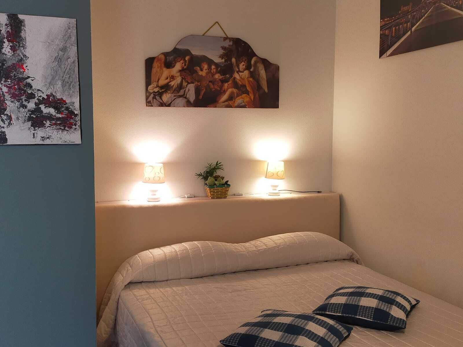 A bedroom with a bed, two bedside lamps, a small plant, and a painting on the wall.