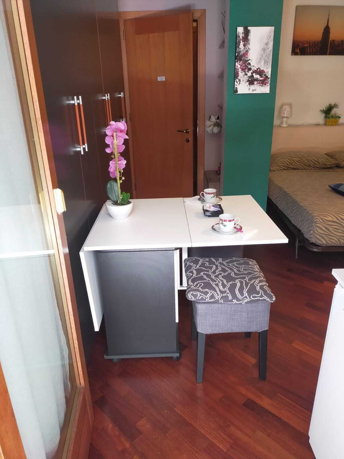 A compact room with a wooden door, a small table with a white tablecloth, a bench, and a bed with a zebra print bedspread.