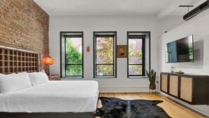 Quarto moderno com parede de tijolos, cama grande, três janelas, TV e planta.