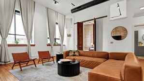 Una sala de estar moderna con un sofá de cuero marrón, sillas de madera, una mesa redonda de centro y un ventanal con cortinas.