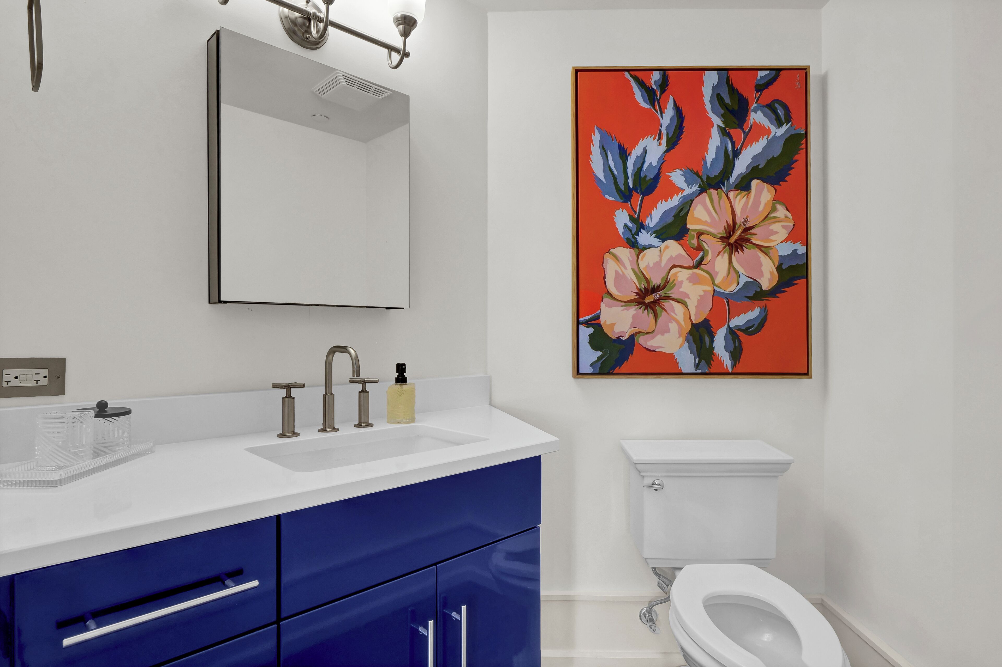 A bathroom with a white sink, blue cabinets, a white toilet, and a large floral painting on the wall.