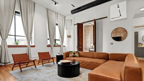Een moderne woonkamer met een bruine leren bank, twee houten stoelen, een ronde zwarte salontafel en een groot raam met gordijnen.
