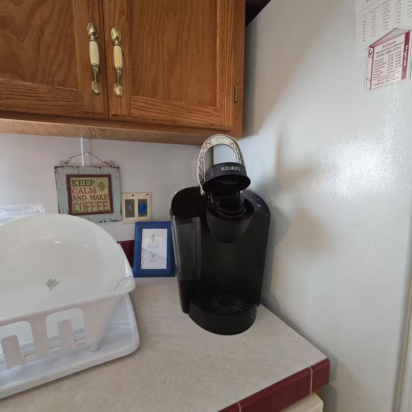 Une cuisine avec des meubles de rangement en bois, une machine à café, un récipient blanc et un panneau encadré accroché au mur.