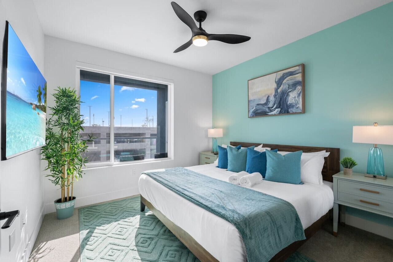 Un dormitorio con cama, ventilador de techo, ventana, planta y un cuadro.