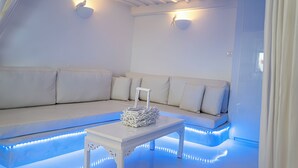 A white room with a sofa, a small table, and a wall-mounted light fixture. - Akrotiri Castle View (Santorini)