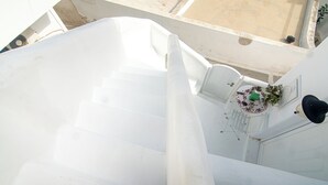 A white staircase leading to a balcony with a table and chairs, a potted plant, and a wall-mounted light fixture. - Akrotiri Castle View (Santorini)