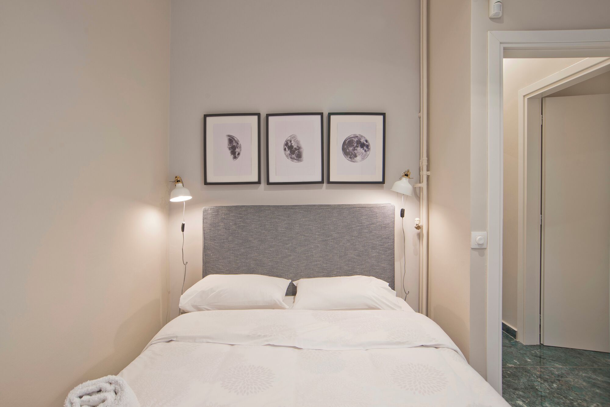 A neatly made bed with white linens, a textured grey headboard, and three framed moon phase pictures on the wall.
