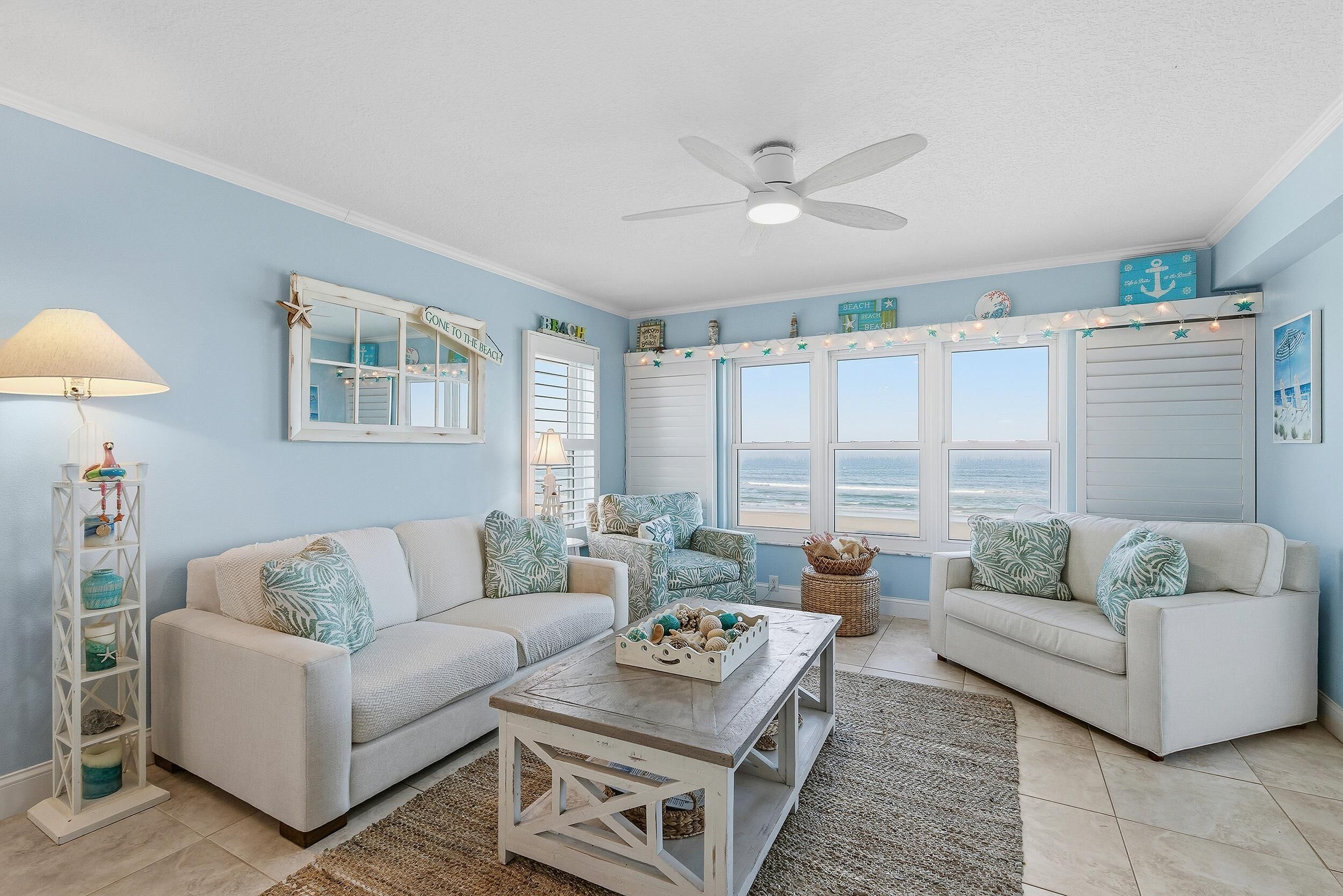 A coastal-themed living room with two sofas, a coffee table, a lamp, and a view of the ocean through large windows.