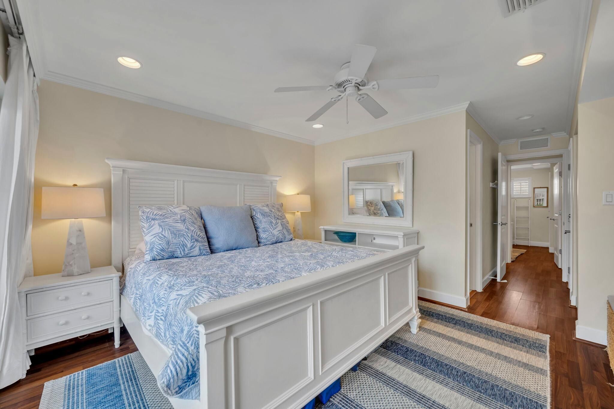 A bedroom with a large bed, white nightstands, a ceiling fan, and a hallway leading to other rooms.