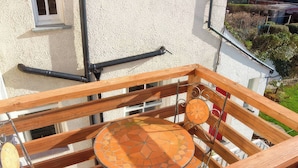 Ein Holzbalkon mit einem Metalltisch und -stühlen, einem kleinen dekorativen Gegenstand und Blick auf ein weißes Gebäude.