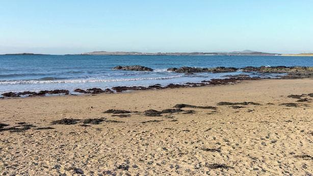 Ein Strand mit felsigem Ufer, blauem Himmel und sanften Hügeln in der Ferne.