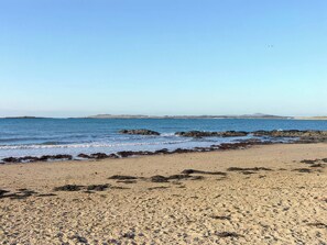 A beach with rocky shoreline, clear blue sky, and distant hills. - Morlais (Rhosneigr)