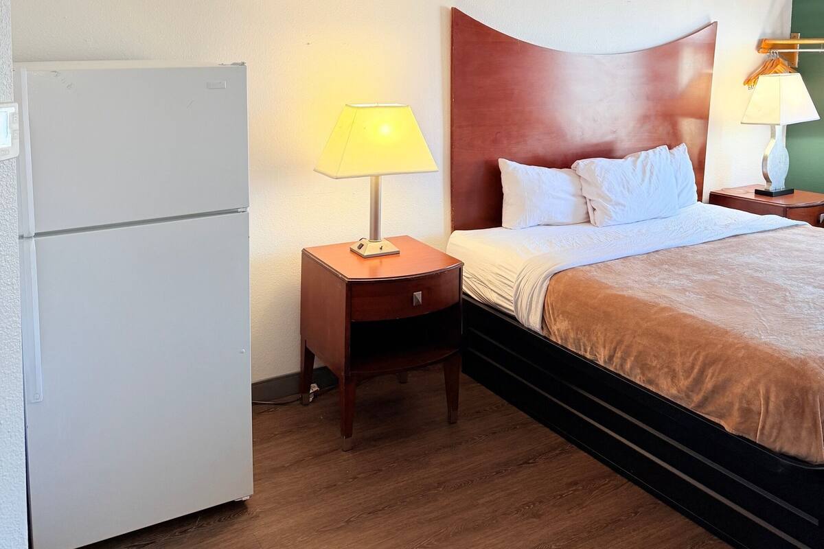 Une chambre d’hôtel avec un lit, une table de chevet avec une lampe, un réfrigérateur et un plancher en bois.