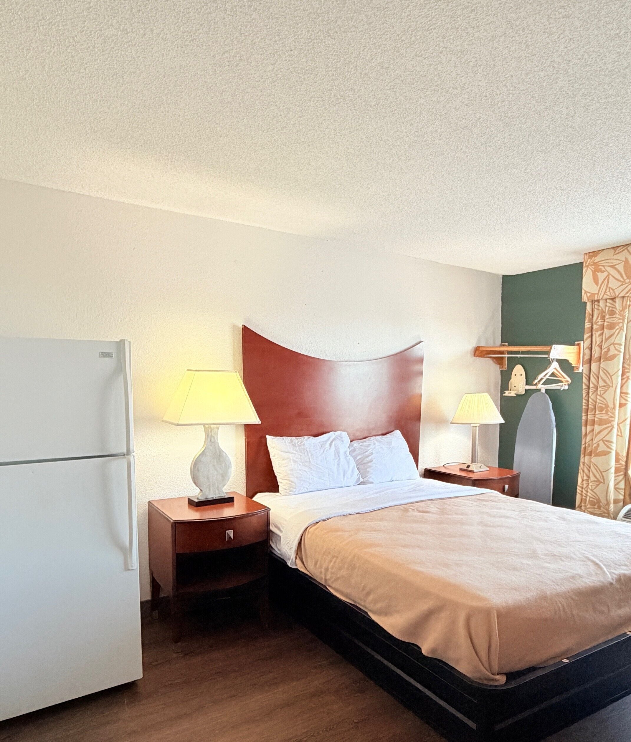 A hotel room with a bed, two bedside tables, a lamp, a mirror, a coat rack, and a refrigerator.