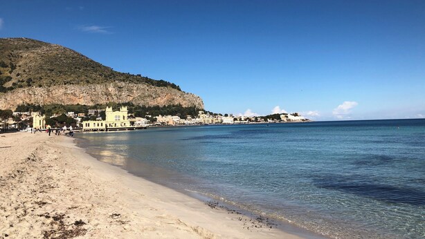 Ein Strand mit klarem Wasser, sandigem Ufer und Gebäuden entlang der Küste.