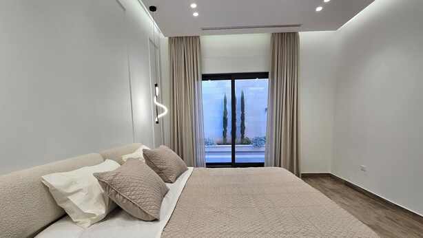 Une chambre à coucher moderne avec un grand lit, une table de chevet et une vue sur les arbres par la fenêtre.