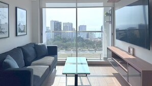 Una sala moderna con sofá, una mesa de vidrio, televisor y balcón con vista a la ciudad.