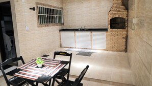 A kitchen with a brick oven, a window with bars, and a table with chairs. - Casa de Praia Pet Friendly com Churrasqueira e Ar (Rio das Ostras)