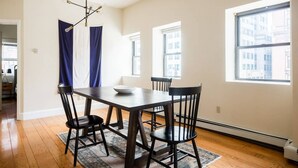 Un coin repas avec une table en bois foncé, quatre chaises noires, un bol blanc et un tapis à motifs. On y trouve deux fenêtres avec des rideaux, et un luminaire est visible au plafond.