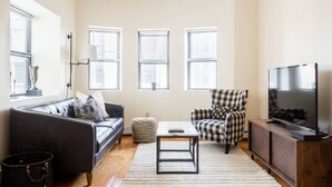 Un salon avec un canapé en cuir noir, un fauteuil à carreaux, une petite table basse, un téléviseur posé sur un meuble en bois et trois fenêtres équipées de stores.