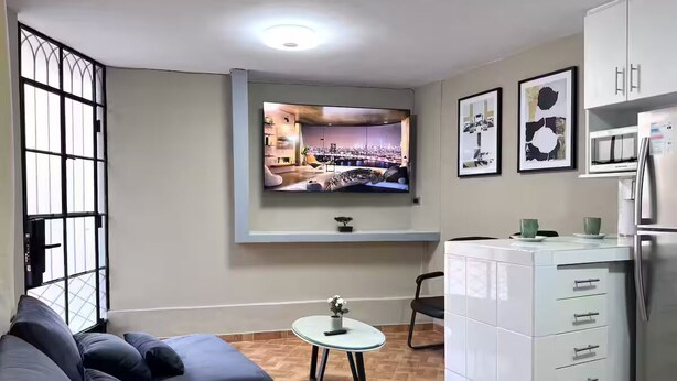 Un ambiente compatto con divano, un piccolo tavolo, televisione a parete e angolo cottura completo di mobili e frigorifero.