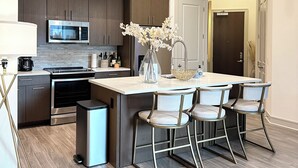 A modern kitchen with dark wood cabinets, a stainless steel microwave, and a large island with bar stools. There are pendant lights above the island and a small trash can to the side. - 1100 Sq Ft Midtown Apt - 2 Miles from Benz Stadium (Atlanta)