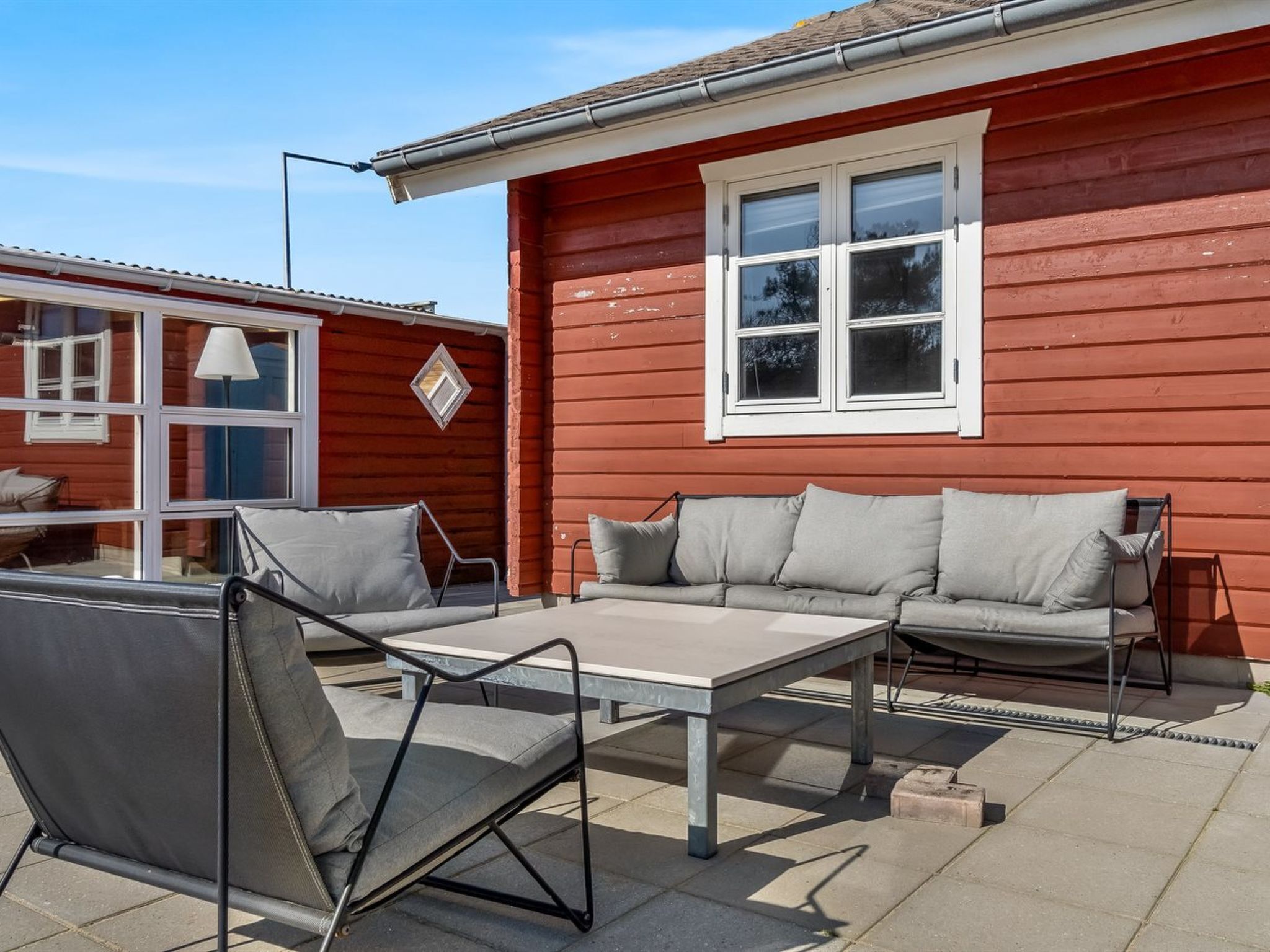 A patio with a metal table and two grey cushioned chairs, a red building with white trim, and a window with white shutters.